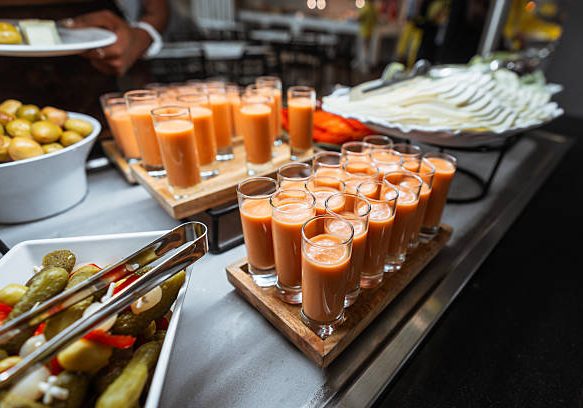 Close-up of a catering table with rows of small glasses filled with chilled orange gazpacho soup, pickles, green olives, and sliced cheese arranged in white dishes; wide-angle indoor shot